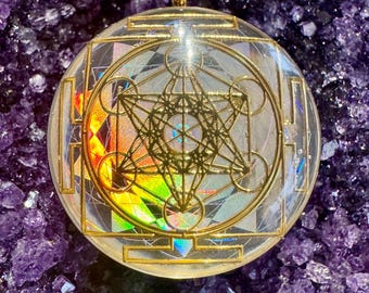 May include: A gold pendant with a clear resin dome featuring a geometric design. The design is a combination of a star of David, a flower of life, and a sacred geometry pattern. The pendant is set against a background of purple amethyst crystals.