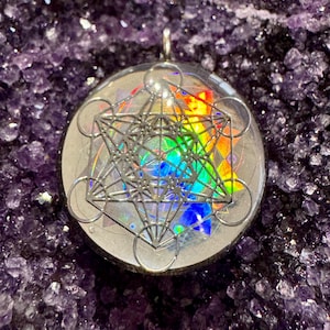 May include: A clear resin pendant with a silver-colored geometric design inside. The design is a combination of circles and lines, creating a complex and intricate pattern. The pendant is set against a background of iridescent rainbow colors.