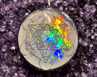 May include: A clear resin pendant with a silver-colored geometric design inside. The design is a combination of circles and lines, creating a complex and intricate pattern. The pendant is set against a background of iridescent rainbow colors.