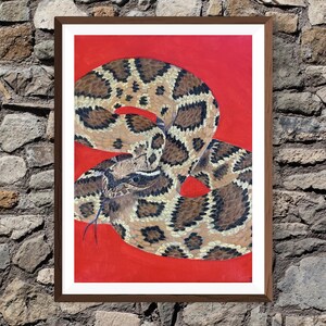 Peut inclure: Peinture encadrée d'un serpent à sonnettes enroulé sur fond rouge. Le serpent est représenté avec des marques marron et noires, et sa langue est sortie. L'œuvre est présentée dans un cadre en bois marron, sur un mur de pierre.