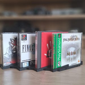 May include: A row of PlayStation game cases, including Final Fantasy VII and Final Fantasy Tactics, displayed in a black plastic holder. The game cases have colorful artwork and text, with the PlayStation logo visible.