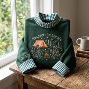 May include: A dark green sweatshirt with a gingham collar and cuffs. The front features an embroidered scene of a tent, a campfire, Bigfoot, and a moth-like creature with the text "Respect the Locals" and "Est. 2026" on the sleeve.