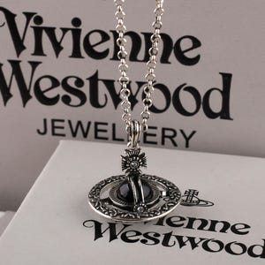 Puede incluir: Un collar de tono plateado con un colgante circular que presenta un anillo exterior intrincado y una esfera central oscura. El colgante está rematado con una pequeña cruz. La cadena consta de eslabones redondos. El logotipo de Vivienne Westwood es visible.