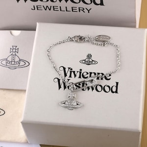 Könnte beinhalten: Ein silberfarbenes Kettenarmband mit einem Sicherheitsnadel-Anhänger und einem zentralen, mit klaren Kristallen besetzten Orb-Anhänger. Der Schmuck liegt auf einer weißen Schachtel mit dem schwarzen Schriftzug Vivienne Westwood und einem Orb-Logo.