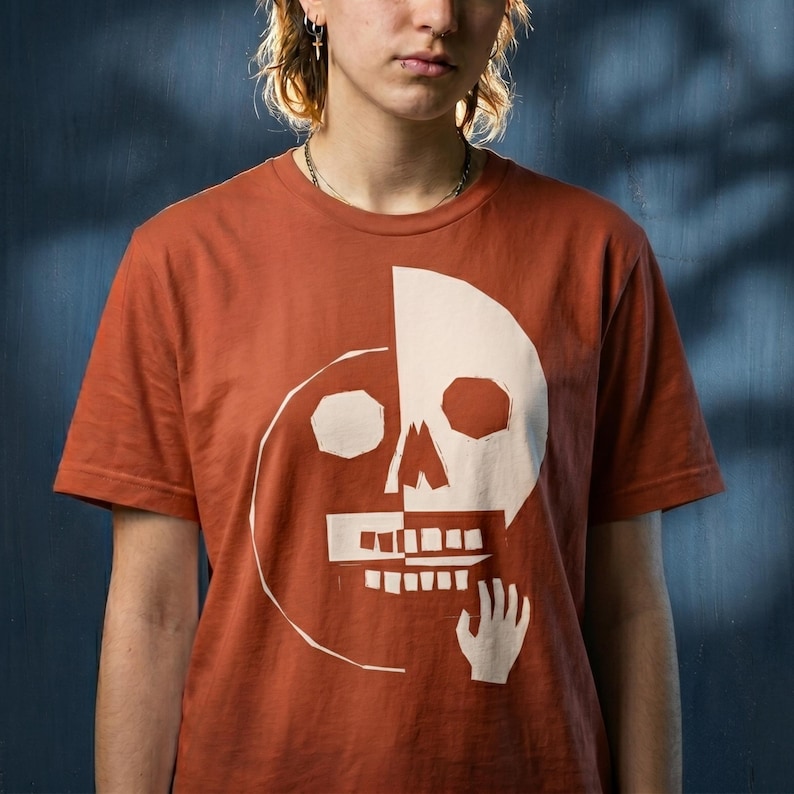 May include: Rust-colored t-shirt featuring a white graphic of a skull divided in half. One side of the skull is complete, while the other side is a simplified outline. A hand graphic is below the jawline. The shirt has short sleeves.
