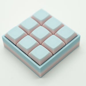 May include: A square, pastel-colored keyboard keycap set. The keycaps are light blue with pink accents, arranged in a 3x3 grid within a matching blue and pink frame. The design is clean and modern.
