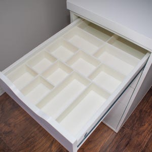 May include: A white drawer pulled open to reveal a built-in organizer with multiple rectangular compartments of varying sizes. The interior of the drawer and the dividers are all finished in a smooth, solid white material, creating a clean look.