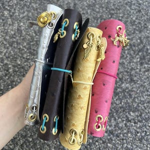 Peut inclure: Un lot de quatre couvertures de journal en cuir argenté, marron foncé, doré et rose. Chaque couverture est ornée d'œillets, de breloques et d'élastiques dorés. La couverture argentée a un charme de chapeau de cow-boy.