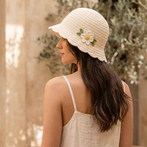 May include: A cream-coloured, woven sun hat with a scalloped brim and a decorative flower. The hat has a small brim and a rounded crown. The flower is white with a yellow centre and green leaves. The hat is a stylish accessory.