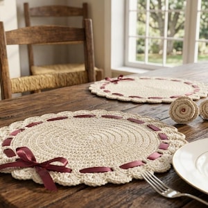 May include: Two round, cream-colored crocheted placemats featuring a scalloped edge and a dark burgundy ribbon woven through the perimeter, tied into a bow on each mat. The placemats are displayed on a wooden table with white plates and forks.