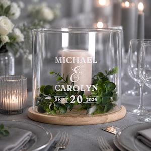 May include: A clear cylindrical glass hurricane candle holder on a round wooden base, containing a beige pillar candle and green foliage. The glass is etched with the text MICHAEL & CAROLYN SEPT 20 2024 in white lettering.