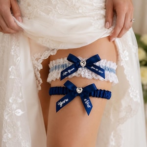 May include: Two wedding garters featuring navy blue satin ribbons, white lace trim, and rhinestone heart embellishments. The top garter displays the text Mrs. Brown and June 20, 2024, while the bottom garter features the text You're Next on the ribbons.