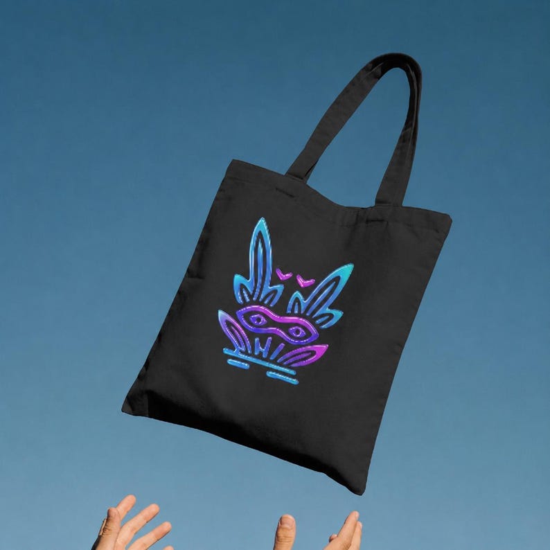 May include: A black tote bag featuring a central graphic design of a stylized creature with large ears and a mask. The artwork uses a vibrant gradient of blue, purple, and pink colors, creating a glowing effect against the dark fabric of the bag.