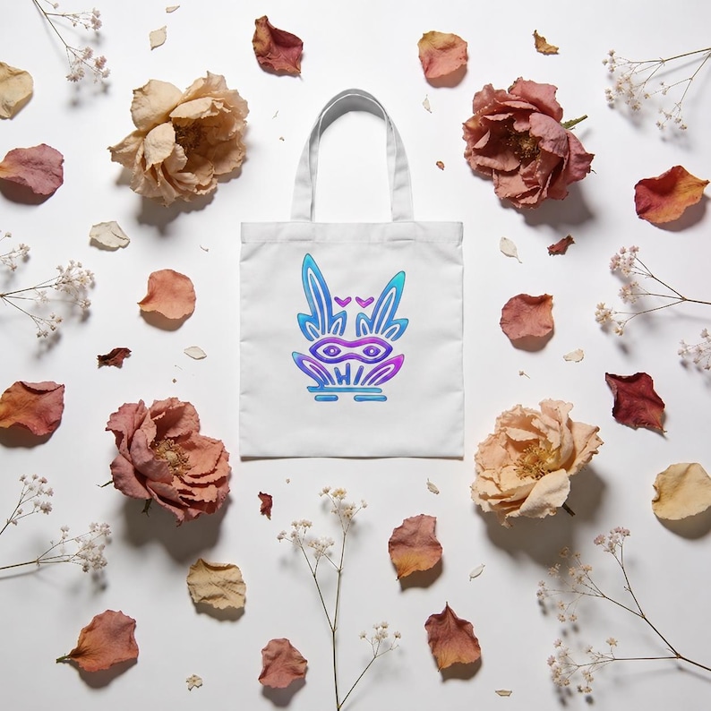 May include: A white tote bag featuring a centered graphic of a stylized creature with long ears, a mask-like face, and a vibrant gradient color scheme transitioning from blue to purple, surrounded by scattered dried rose petals and small white flowers.