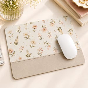 May include: A rectangular mouse pad featuring a cream-colored surface with a repeating pattern of soft pink and peach flowers with green leaves, complemented by a solid beige wrist rest section at the bottom and a white computer mouse on top.