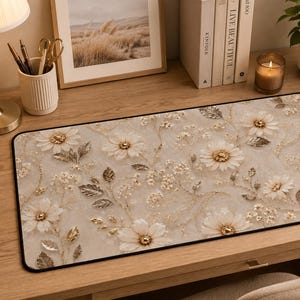 May include: A rectangular desk mat featuring a repeating pattern of white daisy-like flowers and metallic gold leaves on a light beige background. The mat has a thin black border around the entire edge, providing a clean finish to the floral design.