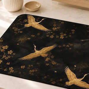 May include: A rectangular desk mat featuring a black background with a repeating pattern of gold-colored flying cranes and small floral blossoms. The mat has rounded corners and a smooth surface, designed to cover a large portion of a workspace.