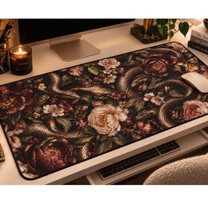May include: A rectangular desk mat featuring a dark, moody botanical print with large, blooming peonies in shades of deep red, pink, and cream, intertwined with coiled snakes and lush green foliage against a black background.