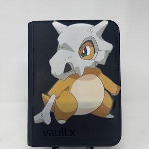 Pode incluir: Um fichário de cartas preto com uma ilustração do personagem Cubone. Cubone tem um capacete de osso e está em tons de bege, marrom e branco. A palavra "vaultx" é impressa em branco na parte inferior.