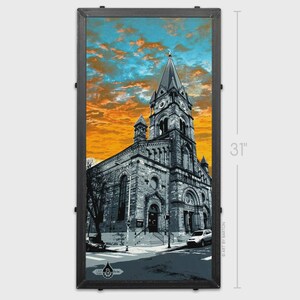 May include: Framed print of a church with a tall steeple, set against a vibrant orange and blue sky. The artwork is 31 inches tall and features a grayscale rendering of the building. The frame is dark and rectangular.