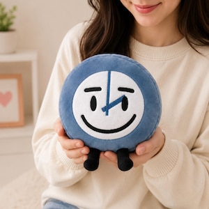 May include: A plush, round, blue clock toy with a smiling face, black feet, and white clock face details. The clock hands are blue. The toy is held in front of a person wearing a cream-colored sweater.