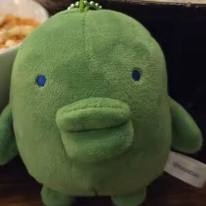 May include: A small, round, green plush toy with a soft, fuzzy texture. It features a prominent, wide, duck-like bill, two small blue circular eyes, and a small green ball chain attached to the top. A white fabric tag with the text BANDAI is visible.