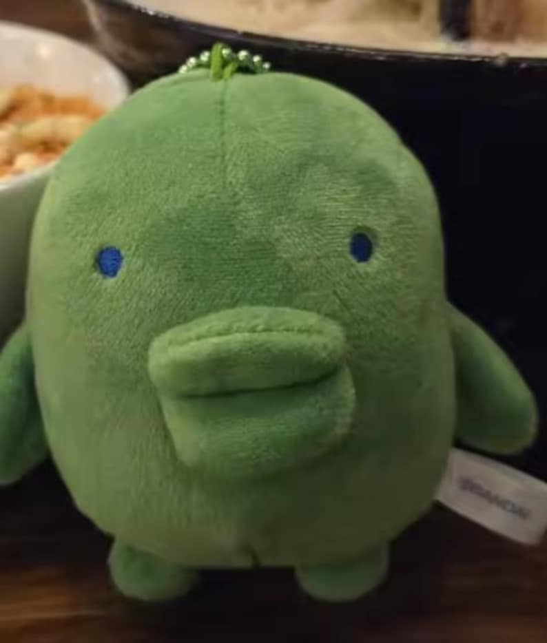 May include: A small, round, green plush toy with a soft, fuzzy texture. It features a prominent, wide, duck-like bill, two small blue circular eyes, and a small green ball chain attached to the top. A white fabric tag with the text BANDAI is visible.