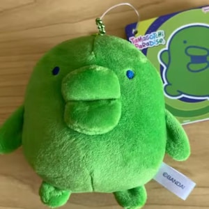 May include: A round, bright green plush toy featuring a wide, flat beak, small blue eyes, and two short arms and legs. A silver ball chain is attached to the top. A tag attached to the side reads BANDAI, and a paper tag displays Tamagotchi Paradise.