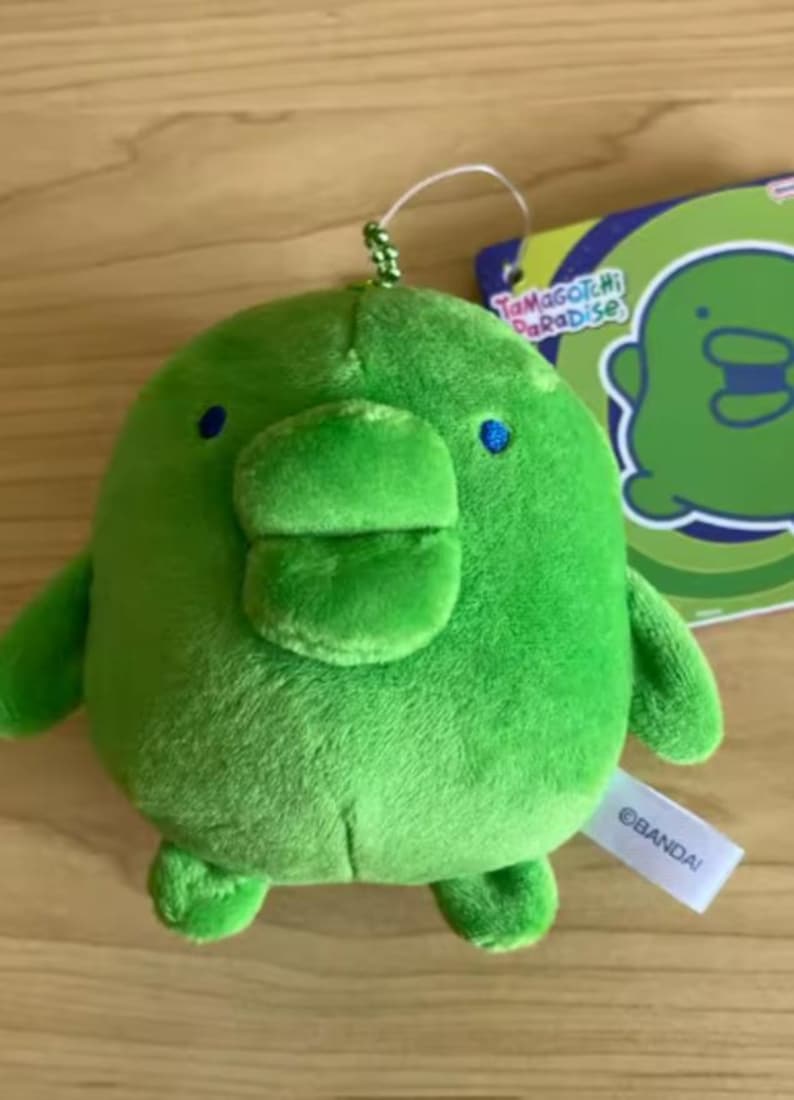 May include: A round, bright green plush toy featuring a wide, flat beak, small blue eyes, and two short arms and legs. A silver ball chain is attached to the top. A tag attached to the side reads BANDAI, and a paper tag displays Tamagotchi Paradise.