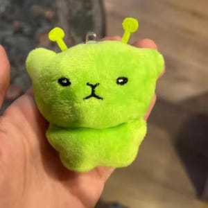 Op de afbeelding: Een limoengroen pluche alien speelgoed met zwart geborduurde ogen en mond. De alien heeft twee gele antennes en een kleine lus om op te hangen. Het speelgoed is zacht en pluizig.
