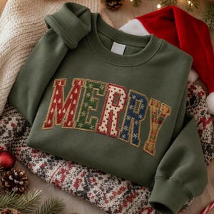 May include: An olive green crewneck sweatshirt featuring the word MERRY in large, block-style letters. Each letter is filled with a different patterned fabric, including red with white trees, green with snowflakes, white with red dots, and blue with stars.