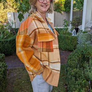May include: A woman wearing a brown, orange and white checked jacket with a floral collar. The jacket has a button-up front and pockets. She is wearing light-coloured trousers.