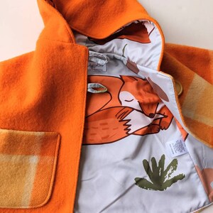 Könnte beinhalten: Orangefarbener Kindermantel mit Kapuze, einer karierten Tasche und einem Fuchs-Musterfutter. Das Futter ist weiß mit orangefarbenen Füchsen und grünen Blättern. Das Etikett lautet "Lullaby Lou" und "Hergestellt in Melbourne".