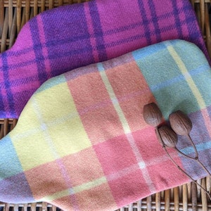 以下が含まれることがあります： 2つの格子柄の布製の湯たんぽカバー。上のカバーは紫とピンクの格子柄。下のカバーは黄色、ピンク、青の格子柄。カバーは籐の編まれた表面の上に置かれている。