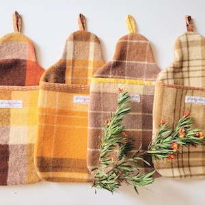 May include: Four rectangular hot water bottle covers made of wool fabric in various plaid patterns of brown, yellow, and cream. Each cover features a top hanging loop and a small white label with the text Lindy Loo sewn on the front pocket.