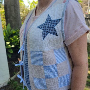May include: A patchwork quilted vest in cream, blue, and beige. A large blue star is sewn onto the front. The vest has blue and white striped trim and blue ties. The vest is worn over a pink top.