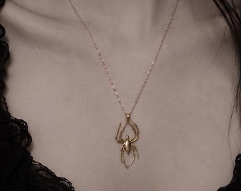 Pode incluir: Um colar com pingente de aranha dourada. O design da aranha é detalhado e pende de uma corrente delicada. O colar é usado em um fundo escuro.