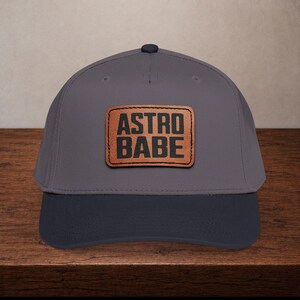 May include: A grey baseball cap with a dark navy blue peak. The cap has a brown leather patch with the words "ASTRO BABE" in bold, black letters. The hat is displayed on a wooden surface.