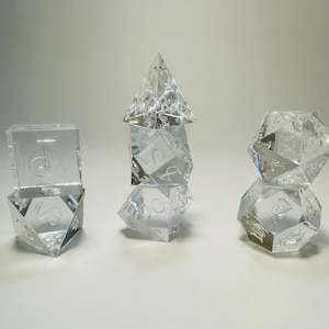 May include: A set of seven clear, transparent polyhedral dice arranged in three stacks. The dice feature engraved numbers and include various geometric shapes such as cubes, pyramids, and multi-faceted spheres used for tabletop gaming.