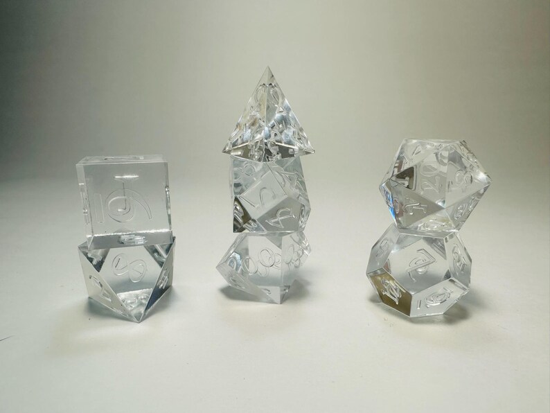 May include: A set of seven clear, transparent polyhedral dice arranged in three stacks. The dice feature engraved numbers and include various geometric shapes such as cubes, pyramids, and multi-faceted spheres used for tabletop gaming.