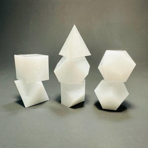 May include: A set of seven translucent white polyhedral dice arranged in three stacks. The dice feature engraved numbers and include a four-sided pyramid, a cube, an octahedron, a dodecahedron, and an icosahedron with various geometric shapes.