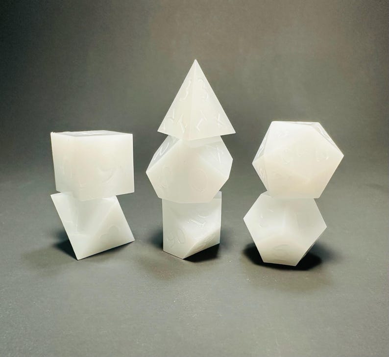 May include: A set of seven translucent white polyhedral dice arranged in three stacks. The dice feature engraved numbers and include a four-sided pyramid, a cube, an octahedron, a dodecahedron, and an icosahedron with various geometric shapes.