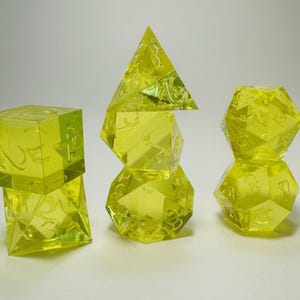 May include: A set of seven translucent neon yellow polyhedral dice arranged in three stacks. The dice feature engraved numbers and include a d4, d6, d8, two d10s, a d12, and a d20, all crafted from a clear, bright yellow resin material.