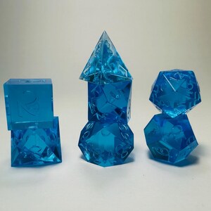 May include: A set of seven translucent blue polyhedral dice arranged in three stacks. The dice feature sharp edges and clear, recessed numbers. The collection includes a d4, d6, d8, d10, d12, d20, and a percentile die, all made of blue resin.