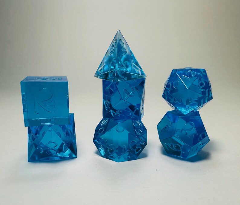 May include: A set of seven translucent blue polyhedral dice arranged in three stacks. The dice feature sharp edges and clear, recessed numbers. The collection includes a d4, d6, d8, d10, d12, d20, and a percentile die, all made of blue resin.