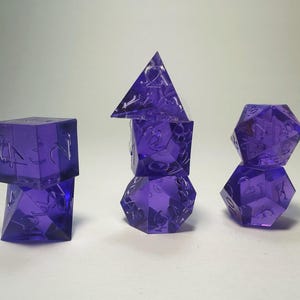 May include: A set of seven translucent purple polyhedral dice arranged in three stacks. The dice feature engraved numbers and include various geometric shapes such as a tetrahedron, cube, octahedron, dodecahedron, and icosahedron for gaming.