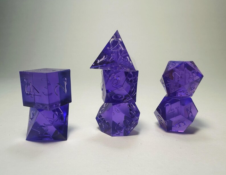 May include: A set of seven translucent purple polyhedral dice arranged in three stacks. The dice feature engraved numbers and include various geometric shapes such as a tetrahedron, cube, octahedron, dodecahedron, and icosahedron for gaming.
