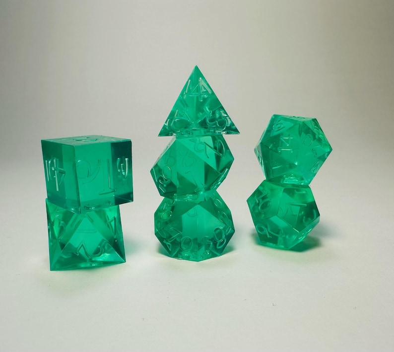 May include: A set of seven translucent emerald green polyhedral dice arranged in three stacks. The dice feature sharp edges and clear, recessed numbers. The collection includes a d4, d6, d8, d10, d12, and d20, all crafted from a smooth, see-through resin material.
