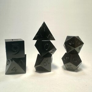 May include: A set of seven black metal polyhedral dice arranged in three stacks. The dice feature engraved numbers in a matching black finish. The collection includes a d4, d6, d8, d10, d12, d20, and a percentile die, all with sharp edges.