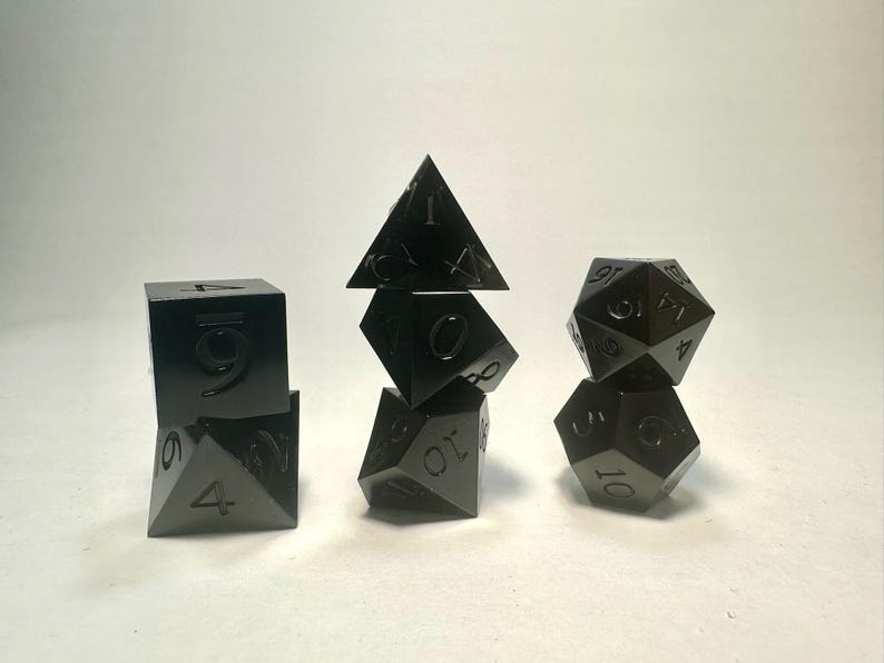 May include: A set of seven black metal polyhedral dice arranged in three stacks. The dice feature engraved numbers in a matching black finish. The collection includes a d4, d6, d8, d10, d12, d20, and a percentile die, all with sharp edges.